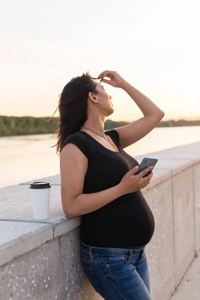 Gravid latinamerikansk kvinna håller smartphone och koppla av på vall med varmt ljus på morgonen — Stockfoto