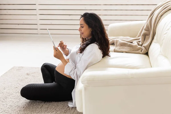 Latin terhes nő táblagéppel ül közel kanapé otthon. Terhesség és tájékoztatás a szülőség koncepciójához. — Stock Fotó