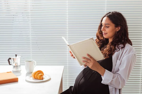 Gravid kvinna äter och läser bok hemma. Morgon, frukost och graviditet koncept — Stockfoto