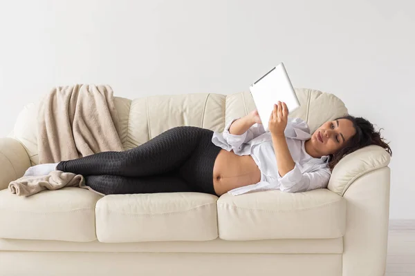 Femme enceinte hispanique utilisant un ordinateur tablette alors qu'elle était allongée sur le canapé à la maison. Grossesse et information pour le concept de parentalité. — Photo