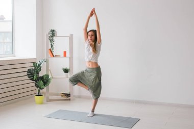 Yoga yapan bir kadın. Saf bir odanın içinde bitkiler ve yeşilliklerle birlikte ağaç duruşu. Sağlıklı yaşam tarzı kavramı. Boşluğu kopyala.