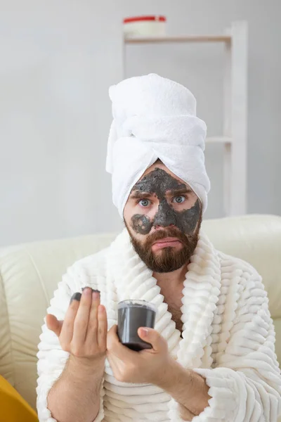 Homem barbudo engraçado se divertindo com uma máscara cosmética em seu rosto feita de barro preto. Homens cuidados com a pele, humor e spa em casa conceito — Fotografia de Stock