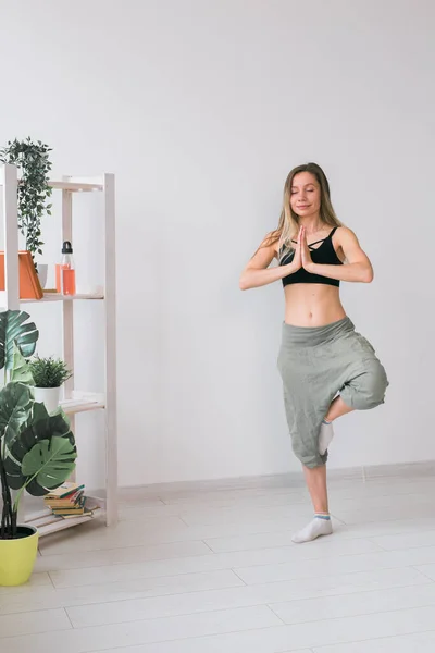 La mujer practica yoga. Árbol pose de pie en la estera en el interior de la habitación acogedora con plantas y vegetación. concepto de estilo de vida saludable — Foto de Stock