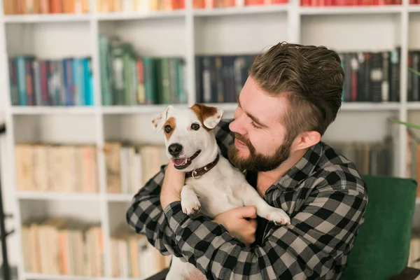 Happy male owner of jack russell terrier dog, feels responsibility of caring about pet, standing against bookshelves background. People and relationship with animals — Stock Photo, Image