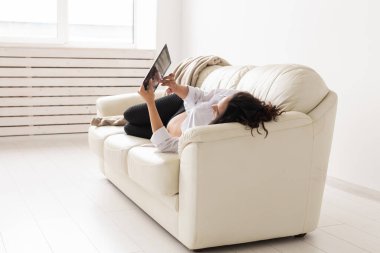 Evdeki kanepede yatarken tablet bilgisayar kullanan İspanyol hamile kadın. Ebeveynlik kavramı için hamilelik ve bilgi.