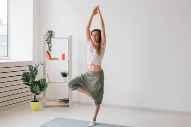 Yoga yapan bir kadın. Saf bir odanın içinde bitkiler ve yeşilliklerle birlikte ağaç duruşu. Sağlıklı yaşam tarzı kavramı. Boşluğu kopyala.