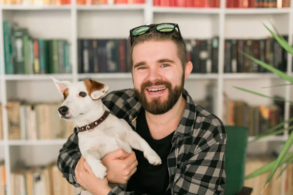 O proprietário masculino feliz do cão terrier de Jack Russell, sente a responsabilidade de importar-se com o animal de estimação, estando contra o fundo de estantes. Pessoas e relacionamento com animais — Fotografia de Stock