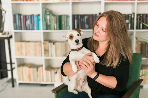 Feliz mujer propietaria de jack russell terrier perro, siente la responsabilidad de cuidar de la mascota, de pie contra los estantes de fondo. Personas y relación con animales — Foto de Stock