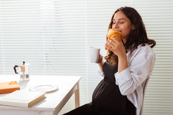 Felice incinta giovane bella donna mangiare croissant durante la colazione del mattino. Concetto di piacevole mattina e atteggiamento positivo durante la gravidanza — Foto Stock