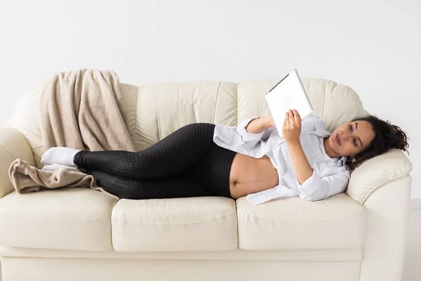 Latin gravid kvinna som använder surfplatta dator när du ligger på soffan hemma. Graviditet och information för föräldraskap koncept. — Stockfoto
