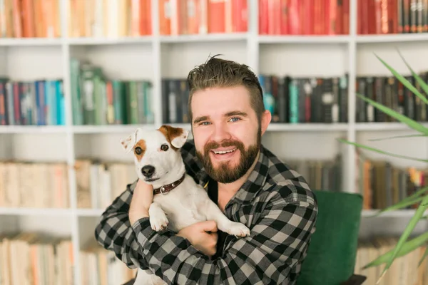 O proprietário masculino feliz do cão terrier de Jack Russell, sente a responsabilidade de importar-se com o animal de estimação, estando contra o fundo de estantes. Pessoas e relacionamento com animais — Fotografia de Stock