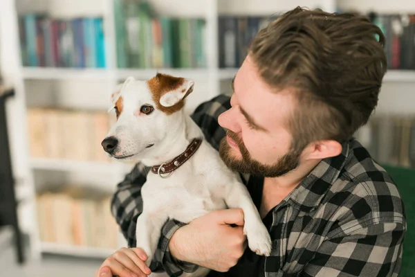 O proprietário masculino feliz do cão terrier de Jack Russell, sente a responsabilidade de importar-se com o animal de estimação, estando contra o fundo de estantes. Pessoas e relacionamento com animais — Fotografia de Stock