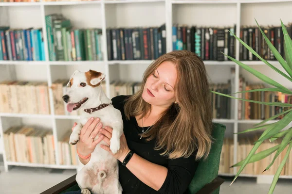 ジャックラッセルテリア犬の幸せな女性の所有者は、本棚の背景に対して立って、ペットの世話の責任を感じている。人と動物との関係 — ストック写真
