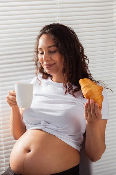 Счастливая беременная красивая женщина ест круассан во время утреннего завтрака. Концепция приятного утра и позитивное отношение во время беременности — стоковое фото