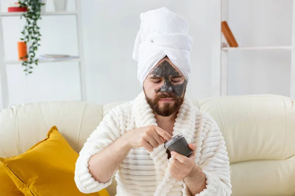 Hombre barbudo divertido divirtiéndose con una máscara cosmética en su cara hecha de arcilla negra. Cuidado de la piel de los hombres, humor y spa en el hogar concepto —  Fotos de Stock