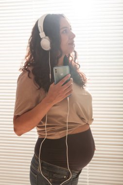 Evdeki pencerenin yanındaki hamile kadının portresi ve kulaklıkla müzik dinlemesi. Hamilelik ve eğlence konsepti.