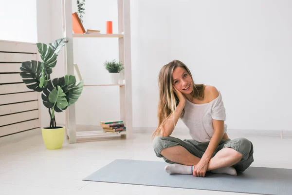 Chica positiva sentada en la colchoneta de fitness descansando después de pilates o práctica de yoga. Concepto de atención plena y bienestar. — Foto de Stock