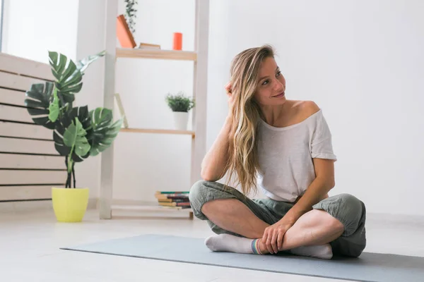 Chica positiva sentada en la colchoneta de fitness descansando después de pilates o práctica de yoga. Concepto de atención plena y bienestar. — Foto de Stock