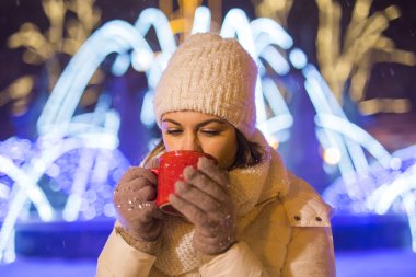 Noel tatili. Kışın sıcak bir fincan kahveyle sıcak kıyafetler içinde gülümseyen güzel bir kadın. Şehir gecesi.