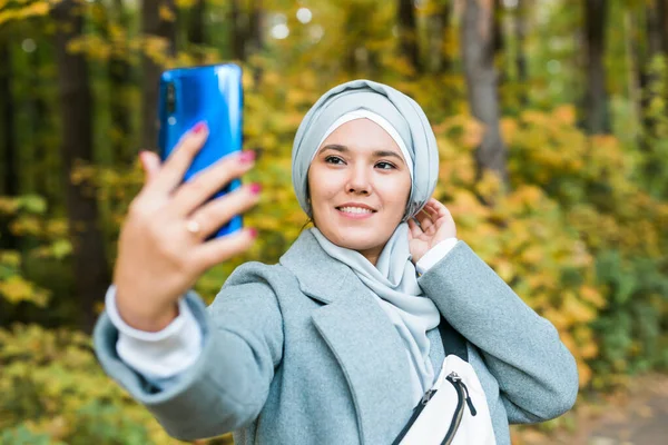 Havalı genç Müslüman Asyalı kız sonbahar parkında açık havada akıllı telefondan selfie çekiyor.. — Stok fotoğraf