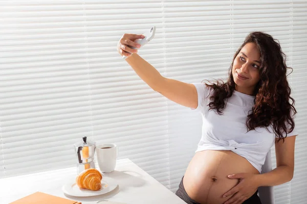 Беременная женщина делает селфи на смартфоне во время завтрака. Концепция декретного отпуска и технологий. — стоковое фото
