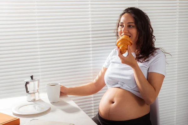 Feliz grávida jovem mulher bonita comer croissant durante o café da manhã. Conceito de manhã agradável e atitude positiva durante a gravidez — Fotografia de Stock