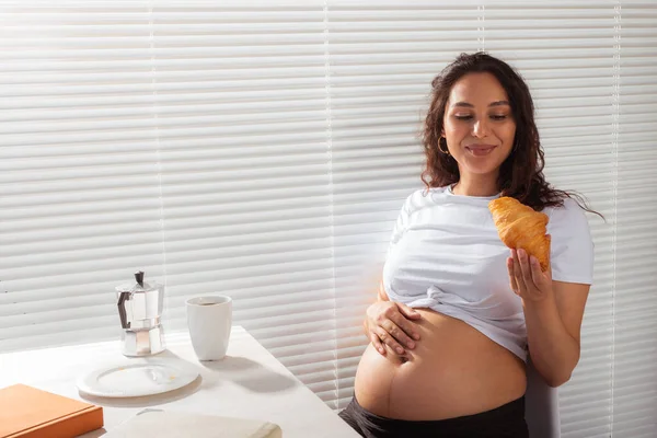 Feliz grávida jovem mulher bonita comer croissant durante o café da manhã. Conceito de manhã agradável e atitude positiva durante a gravidez — Fotografia de Stock