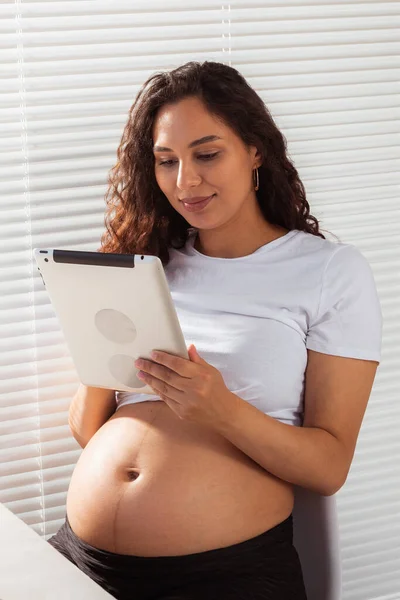 Mujer embarazada hispana usando tableta digital durante el desayuno. Tecnología, embarazo y permiso de maternidad —  Fotos de Stock