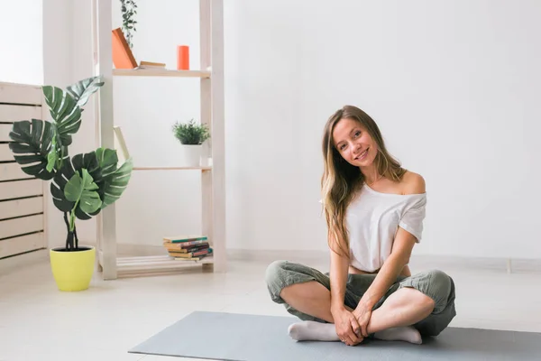 Chica positiva sentada en la colchoneta de fitness descansando después de pilates o práctica de yoga. Concepto de atención plena y bienestar. — Foto de Stock