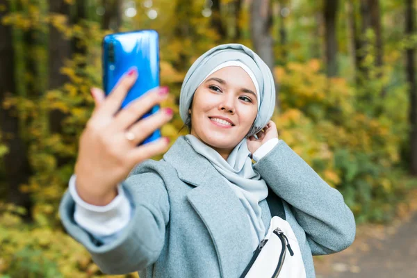 Modische junge muslimische Asiatin im Hidschab, die draußen im Herbstpark ein Selfie mit dem Smartphone macht. Kopierraum. — Stockfoto