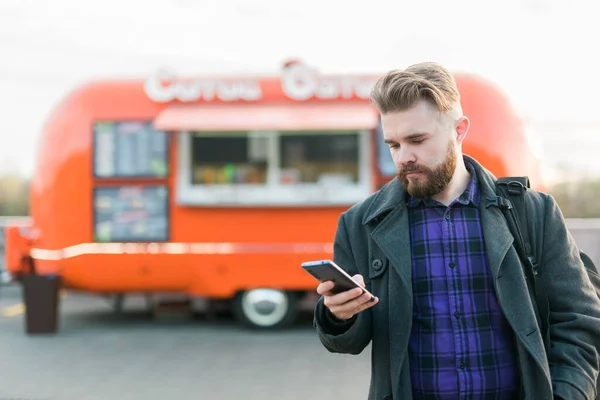 Retrato de joven guapo con teléfono inteligente de pie delante de camión de comida — Foto de Stock