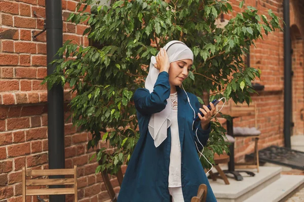 Porträtt av ung modern arabisk kvinna som håller mobiltelefon och lyssnar på musik till hörlurar. — Stockfoto