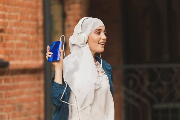 Retrato de jovem árabe moderno mulher segurando telefone celular e ouvir a música para fone de ouvido. — Fotografia de Stock
