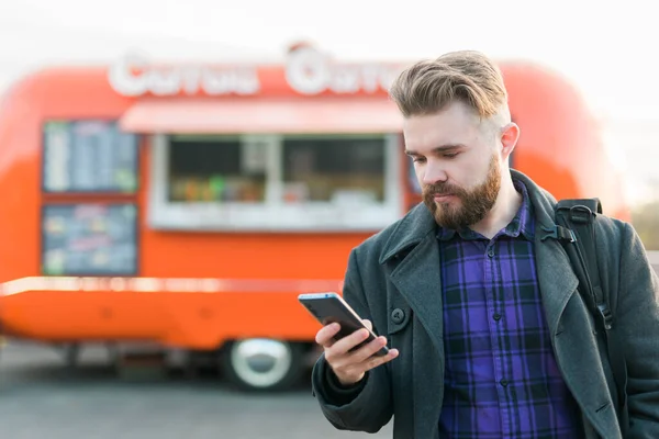 Retrato de joven guapo con teléfono inteligente de pie delante de camión de comida — Foto de Stock