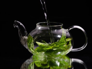 Siyah zemin üzerinde cam bir çaydanlığa sıcak su dökmek, yeşil nane çayı. Bitki çayı ve sağlıklı içecek konsepti.