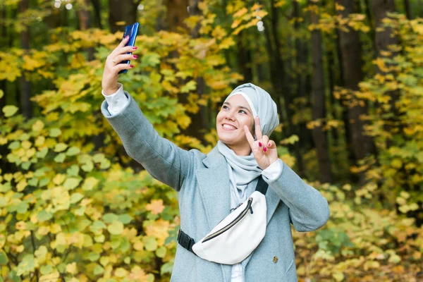 Modische junge muslimische Asiatin im Hidschab, die draußen im Herbstpark ein Selfie mit dem Smartphone macht. Kopierraum. — Stockfoto