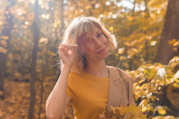 年轻的金发女人用红枫叶遮住一只眼睛.秋天和季节的概念。户外落叶女人像与树叶的特写 — 图库照片