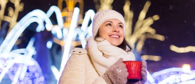 Noel tatili. Kışın sıcak bir fincan kahveyle sıcak kıyafetler içinde gülümseyen güzel bir kadın. Şehir gecesi.