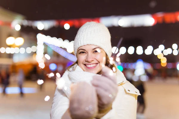 Giovane donna sorridente che indossa vestiti a maglia invernali che tengono scintillante all'aperto su sfondo neve. Vacanze di Natale. — Foto Stock
