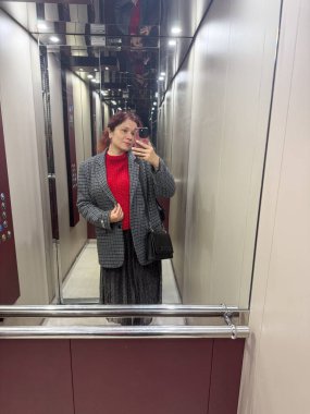 Asansörün içinde şık katmanlı kıyafetlerle selfie çeken bir kadın. Kentsel kadınlık, güven ve kişisel kimlik yansıtıcı modern çevrede
