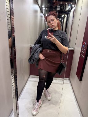 Asansörde aynalı selfie çeken bir kadın siyah tişört, bordo etek ve yırtık tayt giyiyor. Kentsel moda, kusurlar ve kendini ifade etme özgünlüğü ve gelişigüzel yaşam tarzını yansıtıyor