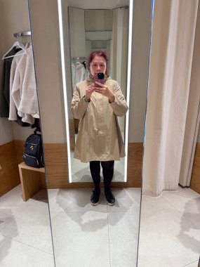 Ayna odasında bej renkli yağmurluk giyen bir kadın. Moda denemesi ve tarafsız kişisel estetik yoluyla kentsel minimalizm, konfor ve öz algı kavramı