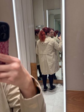 Bej yağmurluk giymiş bir kadının arka ayna yansıması. Siluetin keşfi, vücut farkındalığı ve modern minimalist moda ortamında ince duygular.