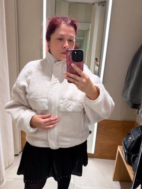 Beyaz yün ceketli kadın ayna fotoğrafı için poz veriyor. Samimi estetiğin, sıcaklığın ve minimalist moda tarzıyla öz farkındalığın temsili