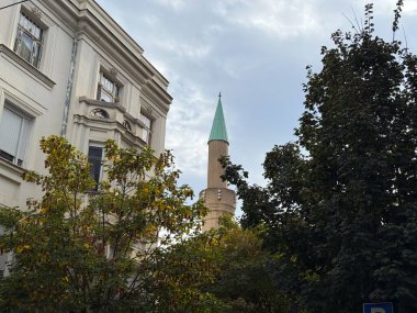 Ağaçlarla çevrili eski beyaz bir binanın yanında yükselen yeşil çatılı uzun bir minare. Din, miras ve birlikte yaşam çeşitli kentsel mimari manzaralar içinde