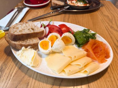 Beyaz tabakta haşlanmış yumurta, ekmek, yeşillik, peynir, somon ve domatesle sabah yemeği. Beslenme ve tazeliği temsil eden dengeli bir kahvaltı.