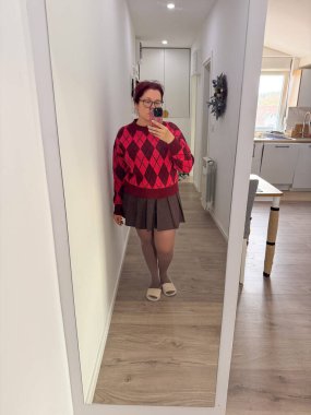 Kırmızı baklava desenli süveter ve kahverengi etekli kadın evde aynalı selfie çekiyor. Rahat kıyafet, sıcak ışık ve sıradan sonbahar tarzı.