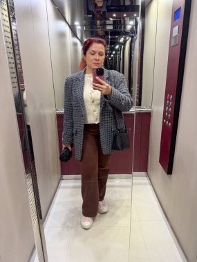Asansörde aynalı selfie çeken bir kadın işaretli ceket ve kahverengi pantolon giyiyor. Sıradan bir anda günlük özgüven, kendini ifade etme ve modern şehir yaşam tarzı