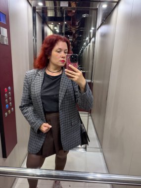 Ceketli ve kısa kahverengi etekli bir kadın asansörde selfie çekiyor. İfade, bireysellik ve şehir güveni günlük hayatın sıradan bir anında yakalanır.