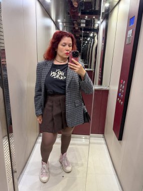 Ceketli ve kısa kahverengi etekli bir kadın asansörde selfie çekiyor. İfade, bireysellik ve şehir güveni günlük hayatın sıradan bir anında yakalanır.